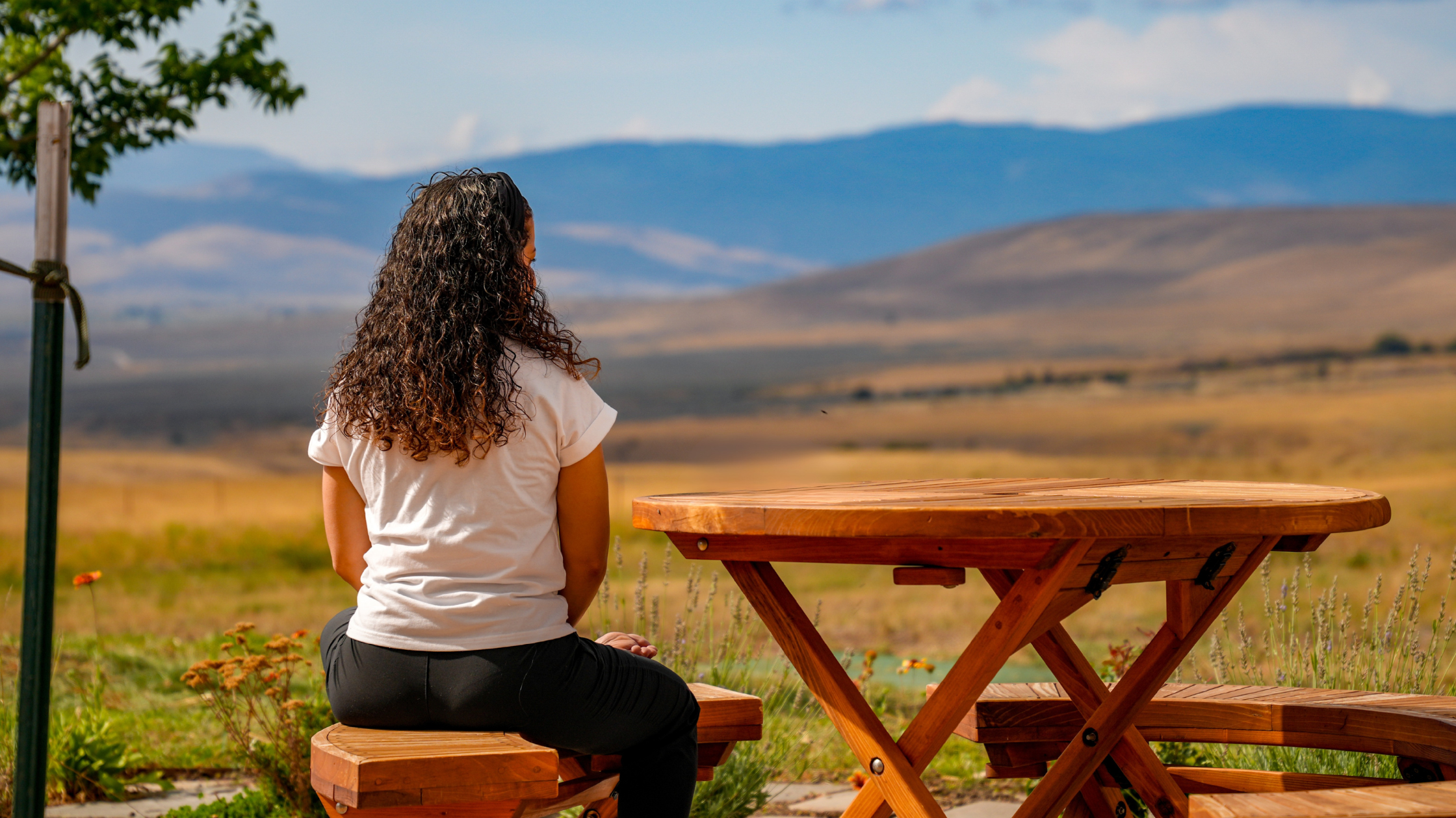 a woman sits on an outdoor picnic table and contemplates the hilly landscape in front of her