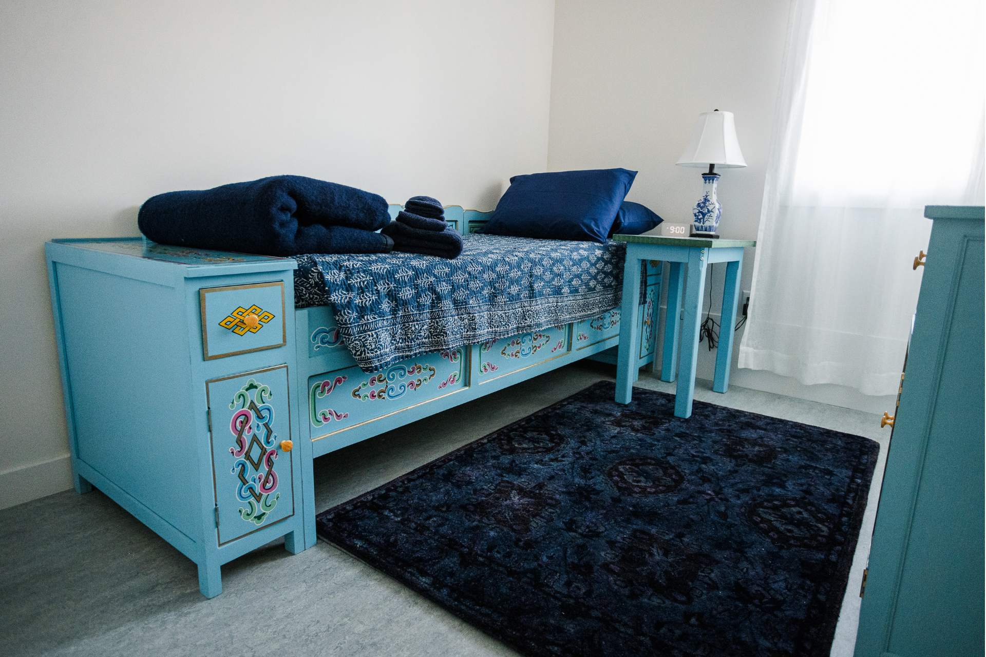 Bedroom A colorful teal bed with dark blue pillows and blankets sits in the corner of a bright, sunlit dorm room.