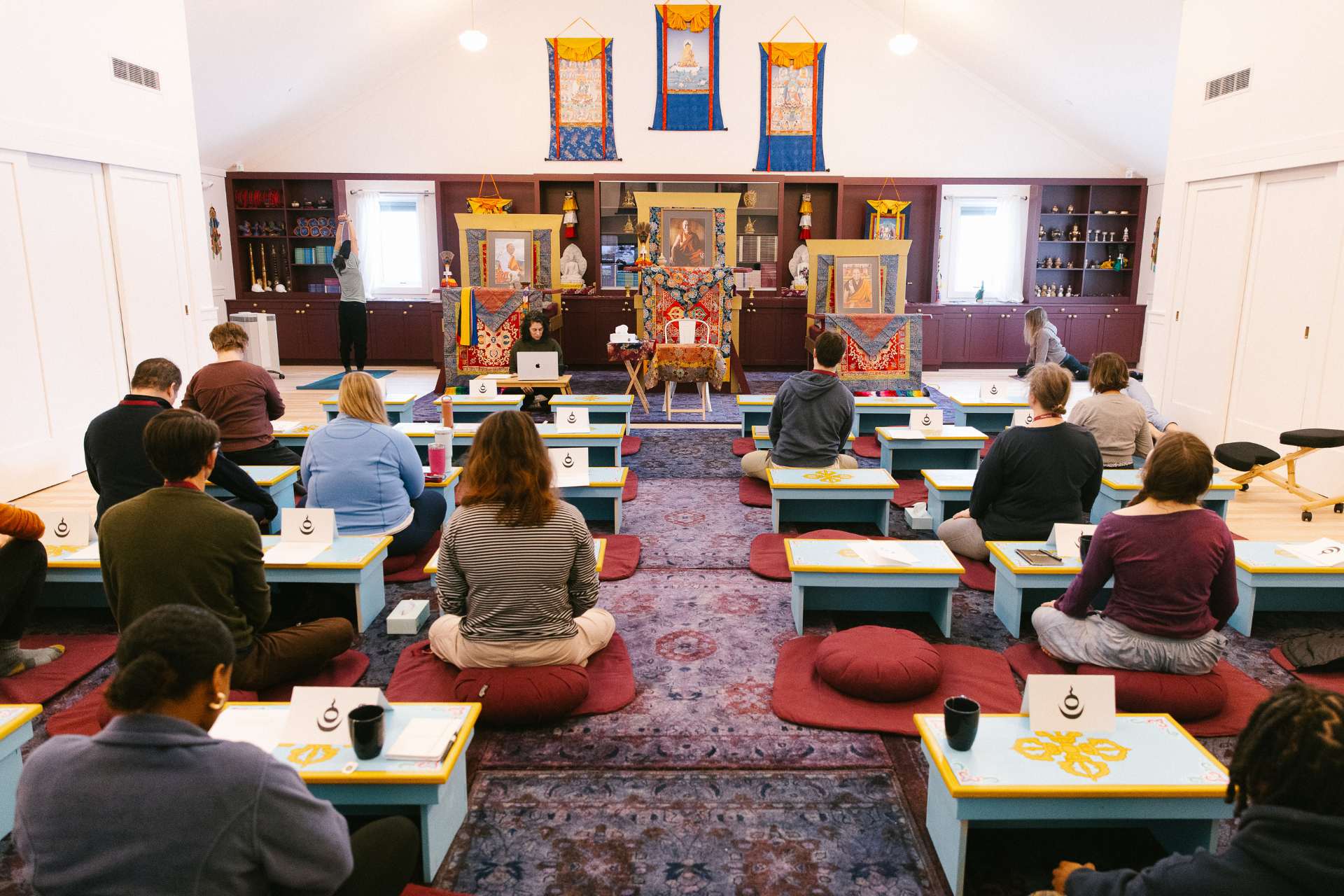 meditation hall A group of students, with their backs to the camera, sit on burgundy cushions in the sunlit meditation hall, listening intently to their instructor.