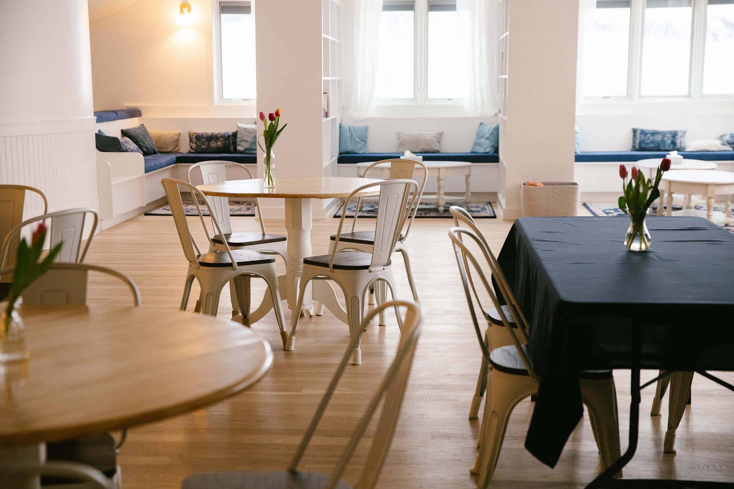 A bright, airy community dining space with white metal chairs, wooden tables, and window seating for retreat guests.