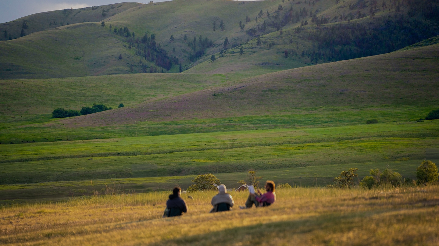 Three people sitting in a grassy field at sunset, engaged in a group reflection during a Namchak retreat.