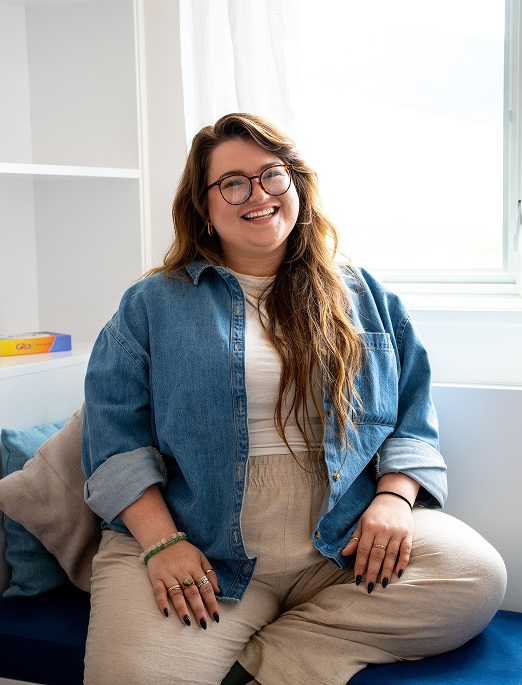 Woman in a blue jean jacket and khaki pants, wearing glasses, smiling while seated on a couch.