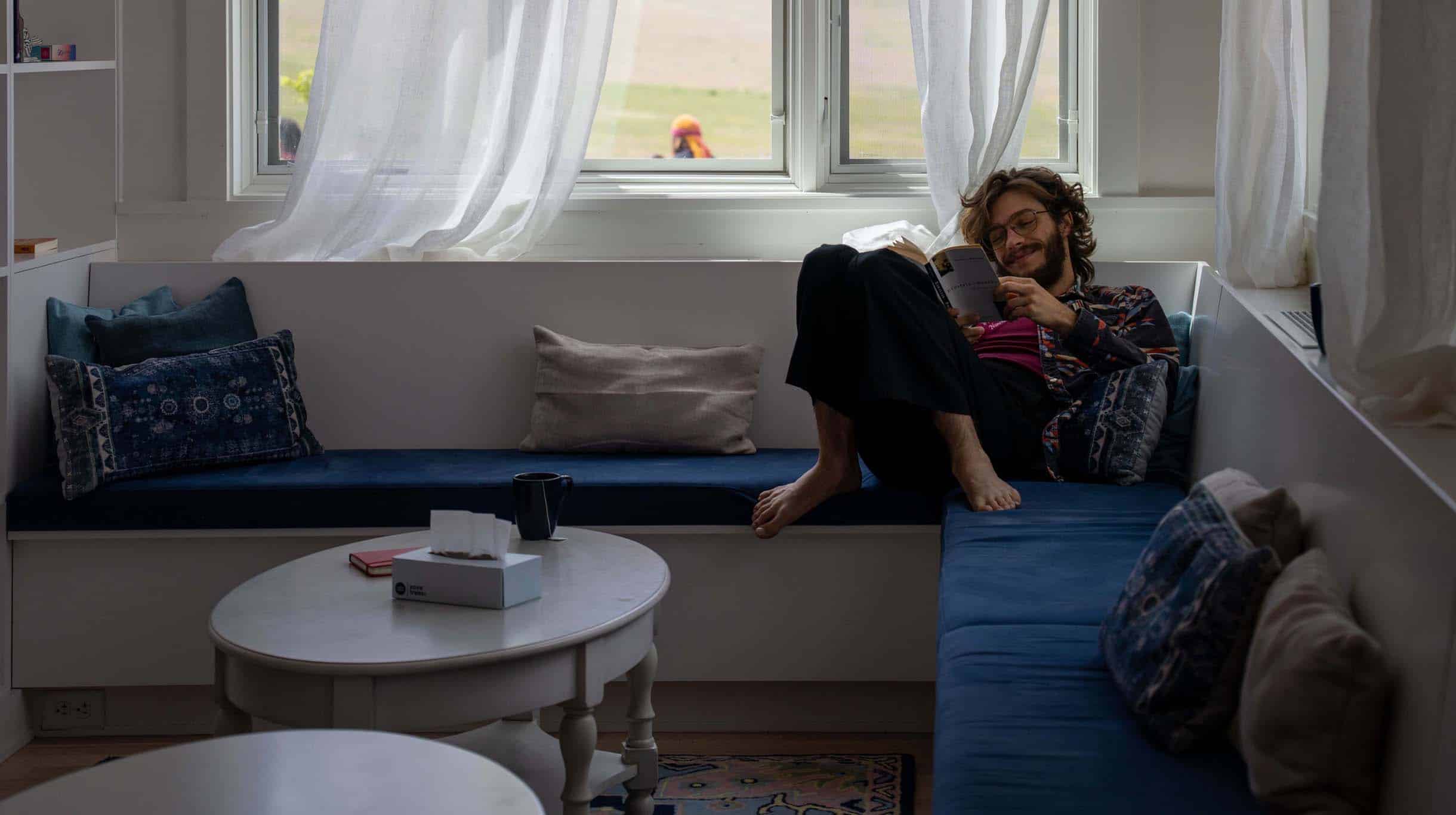 A student lounges on a blue cushioned sofa by a window at the Namchak Retreat Ranch, reading a book.