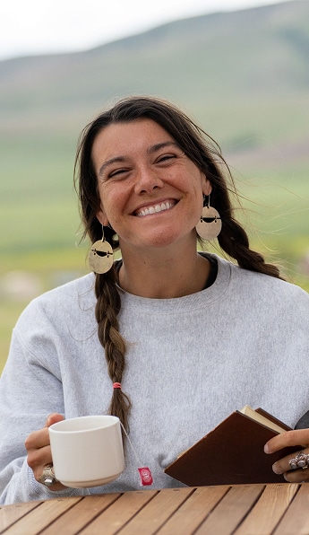 Smiling woman with braids holding a mug and book at an outdoor table at the Namchak Retreat Ranch.