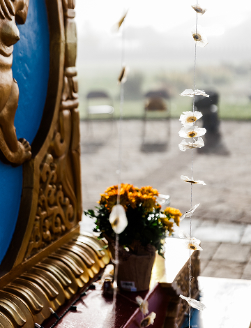 A close-up of a golden Tibetan altar detail and orange marigolds at the Namchak retreat center, with delicate prayer flags hanging in the foreground.
