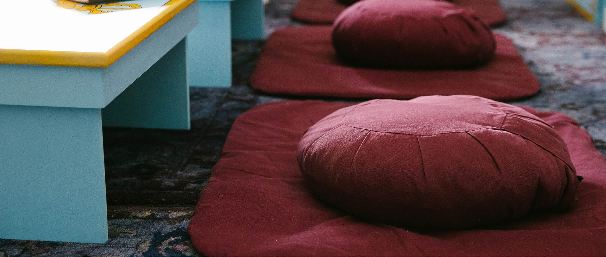 A row of round maroon meditation cushions on matching mats arranged on a floor next to light blue benches at Namchak Retreat Ranch.