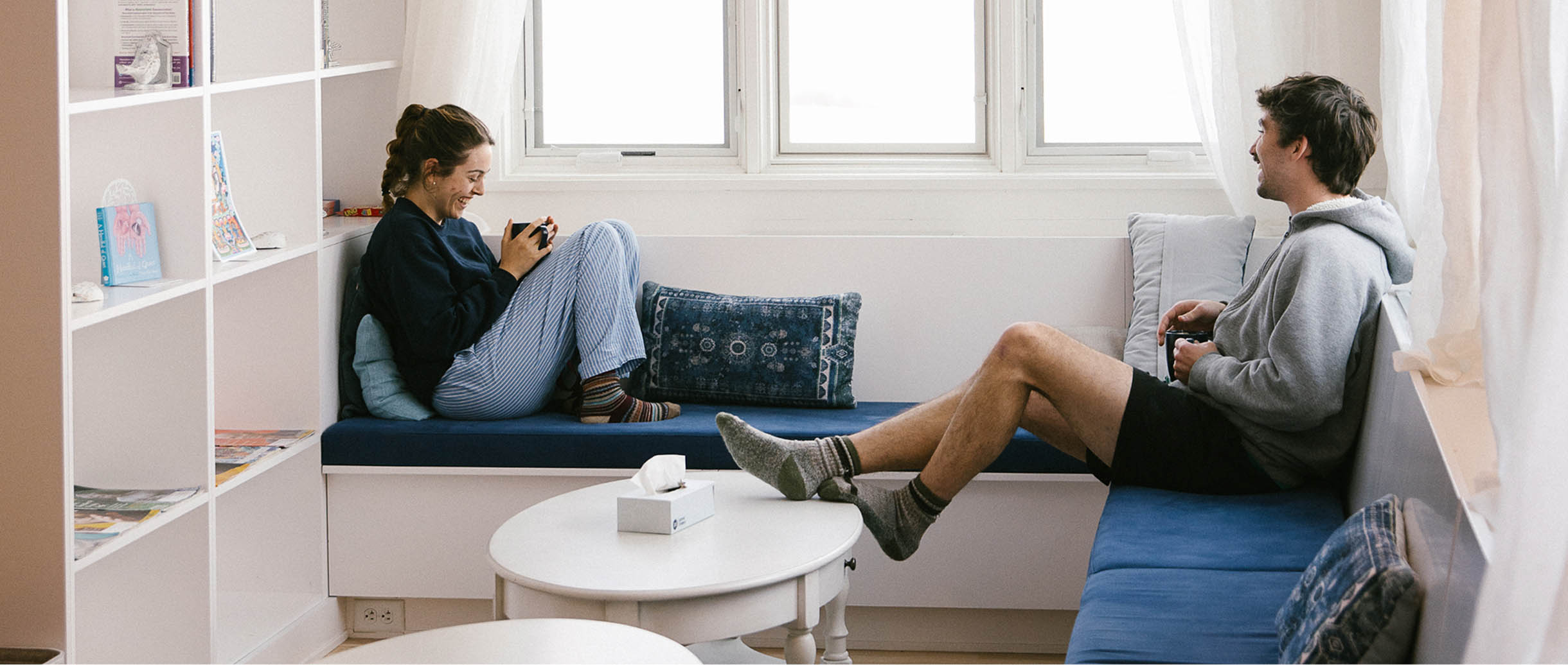 Two students enjoying coffee and conversation in a cozy, modern lounge area with large windows and white bookshelves at the Namchak Retreat Ranch.